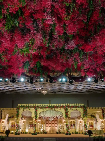 I transform grand ballrooms into royal courts. This wedding stage is defined by its magnificent ceiling, a dense canopy of crimson and green florals that creates an atmosphere of immersive, regal opulence and provides a breathtaking backdrop for the ceremony.