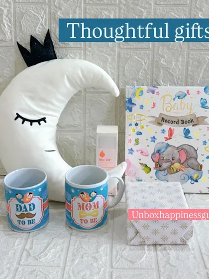 The contents of the moon-themed hamper displayed individually. This set includes "Dad to be" and "Mom to be" mugs, a baby record book, Bio-Oil, a soft cushion, and a wrapped gift.
