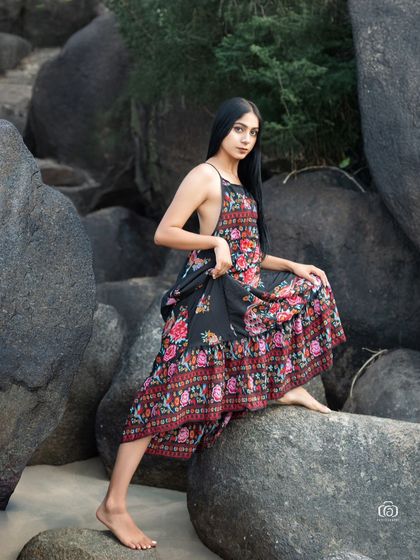 A full-length portrait of the model sitting on a rock, showcasing the flow of the floral dress. The image captures a quiet, peaceful moment by the sea.
