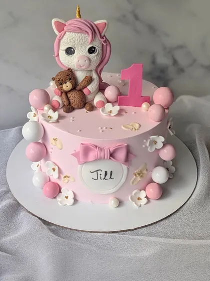 Unleashing magic with this adorable unicorn cake for a first birthday. It features a cute, handcrafted unicorn holding a teddy bear, surrounded by pink and white spheres.
