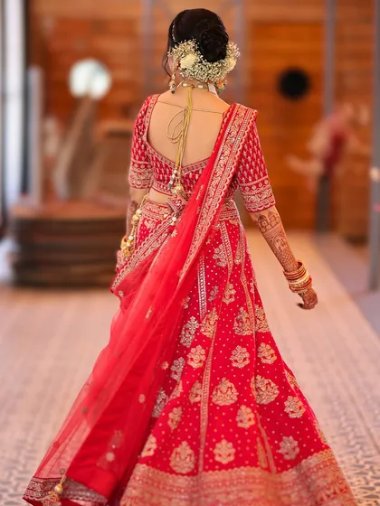 A stunning back shot of the bride. This view highlights the intricate details of her red lehenga and the beautiful floral bun, a classic choice for any bride.