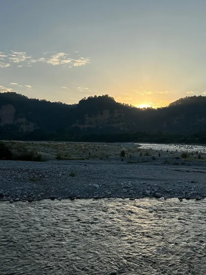 The sun sets behind the hills along a river in Jim Corbett. The gentle light creates a calm and serene mood, reflecting softly on the flowing water.
