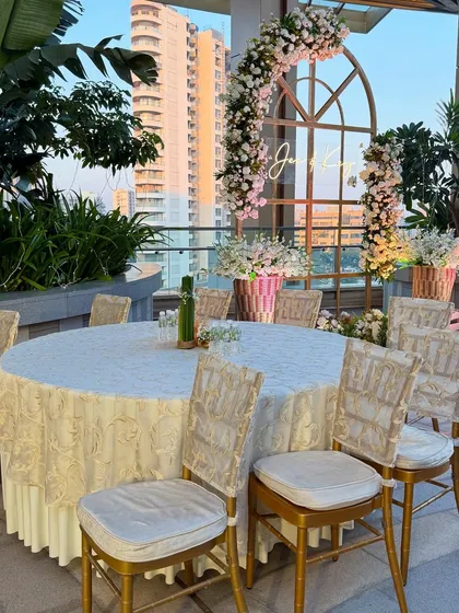 An elegant dining table set for a rooftop celebration. The floral-adorned window arch in the background serves as a beautiful photo-op, framing the city view.