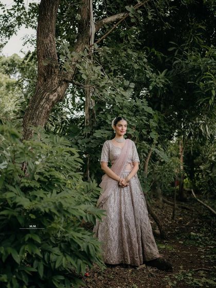 A full-length portrait in a serene, wooded area. The bride stands gracefully, her elegant lehenga contrasting beautifully with the natural surroundings.