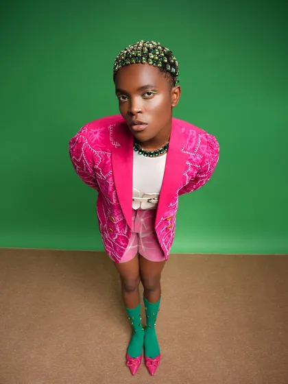 A full view of the bejeweled hair look. The contrast between the vibrant pink jacket and the green gems on the hair and eyes makes the entire image pop.