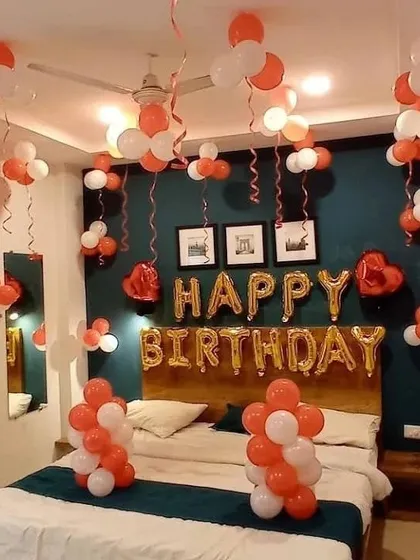 A cheerful birthday room setup with orange and white balloons. The decor includes 'Happy Birthday' foil balloons, balloon clusters on the walls, and balloons with ribbons hanging from the ceiling.