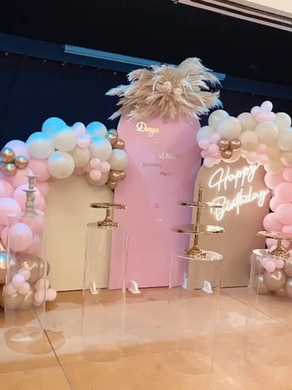 A chic and modern birthday setup in pink and gold. This arrangement features custom-arched backdrops, a neon sign, and clear acrylic pedestals for a clean and sophisticated look.