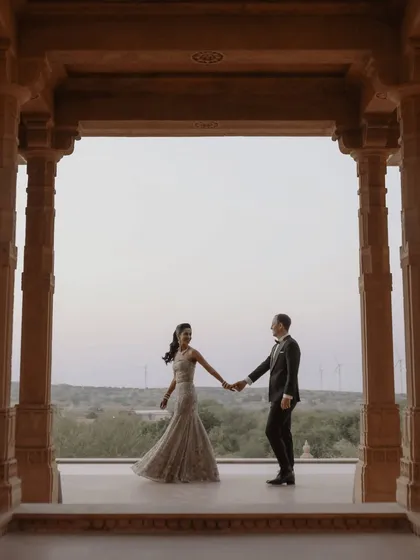 A classic, elegant pose within the pillared corridors of Suryagarh, Jaisalmer. The couple holds hands, their formal attire contrasting beautifully with the ancient stone and the expansive desert landscape visible beyond.