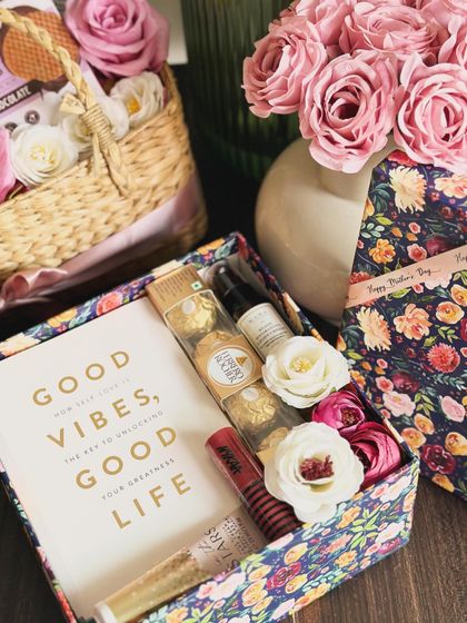 A thoughtful Mother's Day gift featuring the book "Good Vibes, Good Life," a Kama hydrating cream, Ferrero Rocher, a Nykaa lip color, and a Bath & Body Works hand cream, all in a floral printed box.