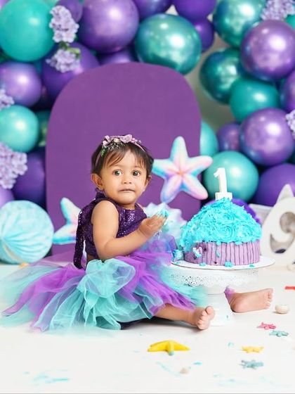 A magical mermaid-themed first birthday cake smash. The purple and teal balloon arrangement and seashell props create an enchanting under-the-sea world for the birthday girl.
