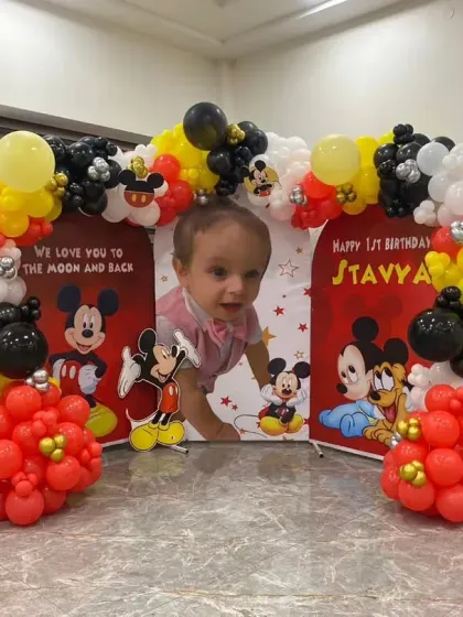 A classic Mickey Mouse theme for a first birthday. This home setup includes a three-panel backdrop with a personalized photo, character cutouts, and a red, yellow, black, and white balloon arch.