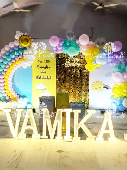 A colorful stage setup for Vamika's "one-derful" first birthday. The decor includes a large balloon rainbow, marquee letters, and a golden sequin panel, creating a bright and joyful atmosphere.