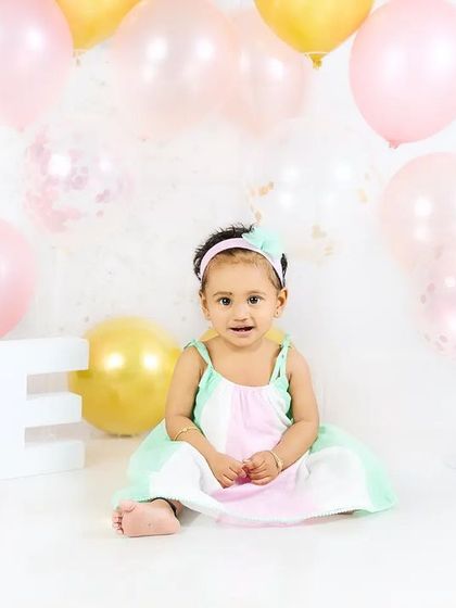 A baby girl in a pastel dress sits in a setup with pink and gold balloons, celebrating her first birthday in style.