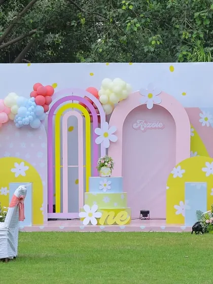 An enchanting outdoor setup for a "Sunshine & Pastel Rainbows" first birthday. This backdrop features whimsical pastel arches, giant balloon daisies, and a custom cake stand, all designed to create a bright and joyful atmosphere in a garden setting.