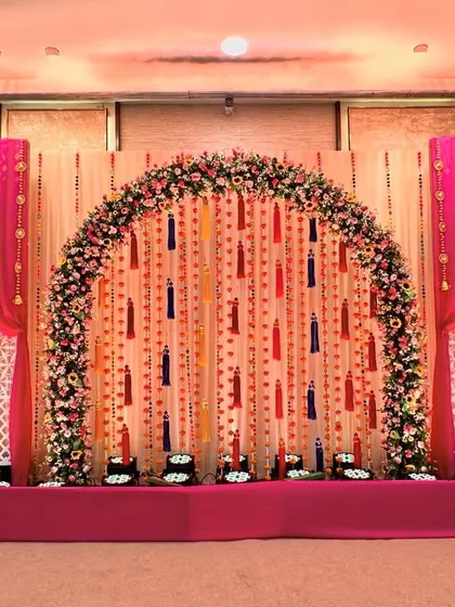 A beautiful Mehendi stage with a floral arch and a backdrop of hanging tassels. The bright pink drapes and lattice panels create a look that is both traditional and celebratory.