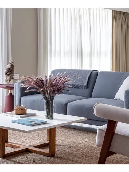 The living room of 'Vishu Home' is designed for comfort and conversation. A soft blue-grey sofa, a white coffee table with a wooden base, and a natural jute rug create a relaxed yet sophisticated atmosphere.