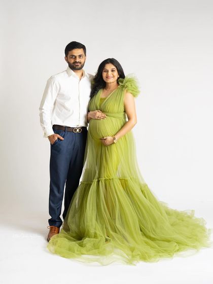 A formal studio portrait of a smiling couple. The mom-to-be wears a beautiful lime green tulle gown, standing next to her partner against a clean white background.