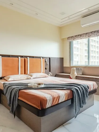 I used a warm, earthy color palette with pops of burnt orange to make this bedroom feel cozy and inviting. The design features a custom upholstered headboard against a fluted wood panel, a comfortable window seat for relaxation, and layered lighting to create a warm and welcoming ambiance.