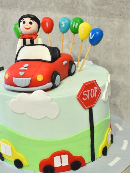 A fun and colorful car-themed cake for a little one's birthday. The cake features a road design, traffic signs, and a cute red car with a driver on top.