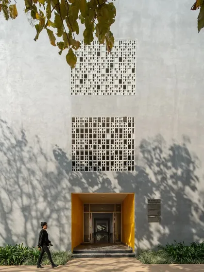 A pedestrian walks past an entrance at Vidyashilp Academy. The deep yellow reveal of the doorway provides a pop of color against the neutral tones of the concrete and 'jaali' facade, acting as a welcoming beacon.