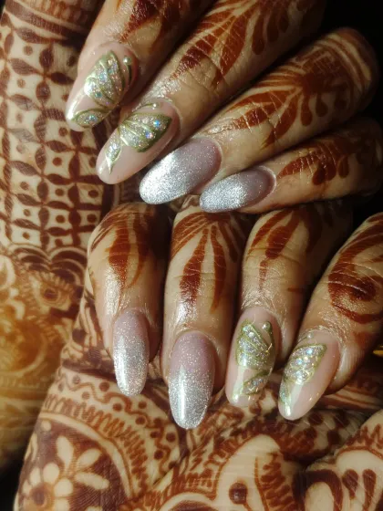 A detailed close-up of bridal nail art. The design includes a shimmering base with hand-painted, glitter-accented leaves that coordinate with the client's henna.