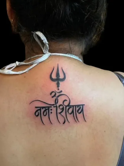 A clean and elegant "Om Namah Shivay" script tattoo on the upper back, topped with a Trishul. This is a classic and powerful mantra tattoo.