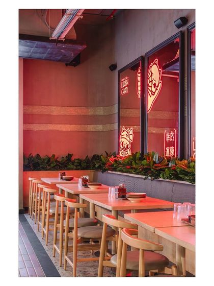 The row of tables along the wall benefits from the ambient red light, creating a cozy and uniform dining section. The simple wood furniture allows the bold lighting and brand elements to stand out.