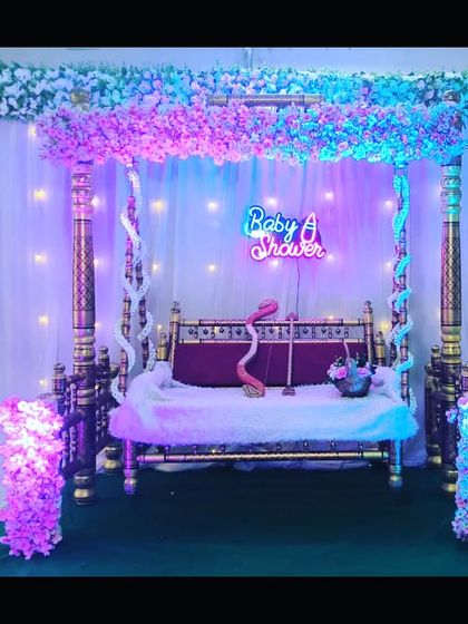 A traditional wooden swing decorated with cherry blossoms and a 'Baby Shower' neon sign. The purple and blue lighting creates a dreamy, enchanting atmosphere.