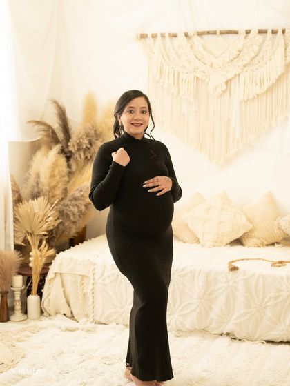 A candid shot of a mother-to-be in a simple black dress, smiling in a cozy boho room. The natural setting creates a relaxed and authentic feel.