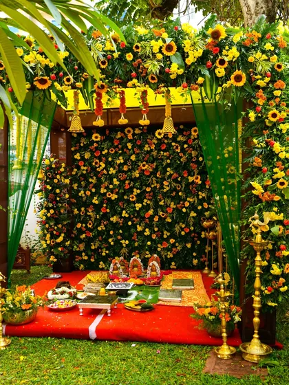 A traditional mandap for an outdoor naming ceremony or housewarming pooja. The structure is covered in a dense arrangement of sunflowers and marigolds, with brass lamps adding to the auspicious feel.