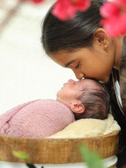 An older sister gives her sleeping newborn sibling a gentle kiss on the forehead. The warm lighting and floral framing make this a particularly sweet sibling portrait.