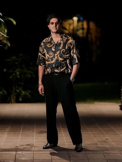 Dulquer Salmaan making a statement in a Versace print shirt for the 'Lucky Bhaskar' promotions in Kochi. Paired with simple black trousers, the bold shirt does all the talking.