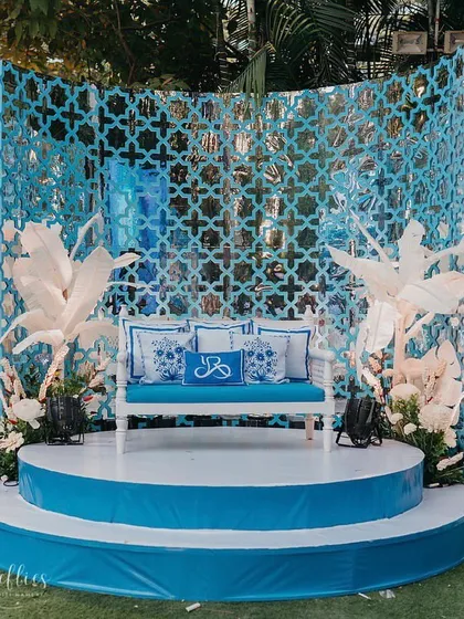 The main stage for the Haldi, featuring a backdrop of intricate blue 'jaali' work and large, handcrafted white paper leaves. The custom-monogrammed cushions on the seating add a personal touch.