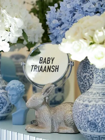 A shelf display with blue and white vases, fresh flowers, and a mirror personalized with 'Baby Triaansh'.