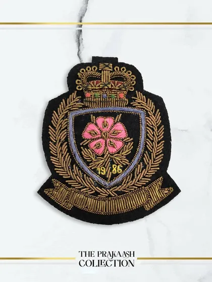 A beautiful hand-embroidered crest patch featuring a pink flower at its center, surrounded by a gold wreath and banner. This detailed appliqué is ideal for personalizing blazers or school-inspired fashion.