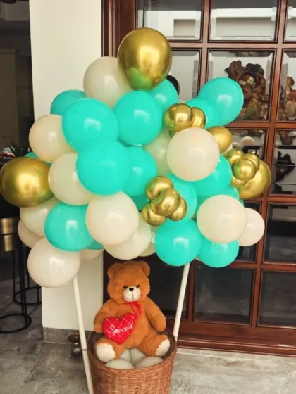 A unique "hot air balloon" decoration for a welcome baby party. The basket holds a cute teddy bear, and the balloon is made from a mix of teal, white, and gold balloons.