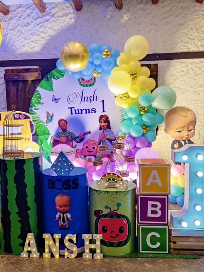 A joint Cocomelon and Boss Baby-themed first birthday. The setup features character cutouts, themed plinths, and a large light-up number '1'.