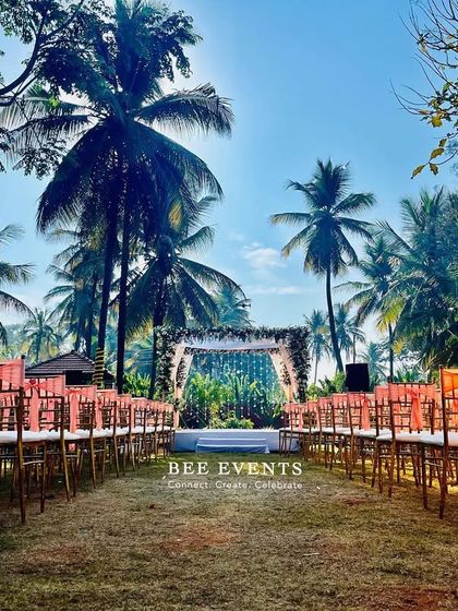 A wide shot of the beautiful outdoor wedding ceremony venue. The aisle is lined with elegant chairs, leading to a stunning floral mantap set against a backdrop of lush greenery and palm trees.