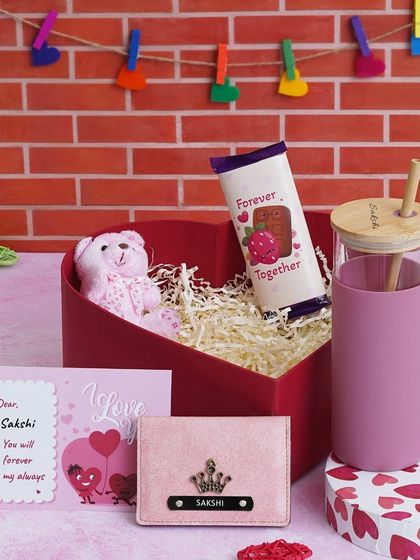 This Love Keepsake Hamper is perfect for her, featuring a personalized glass tumbler, a mini wallet with a custom charm, a teddy bear, chocolate, and a message card, all in a heart-shaped box.
