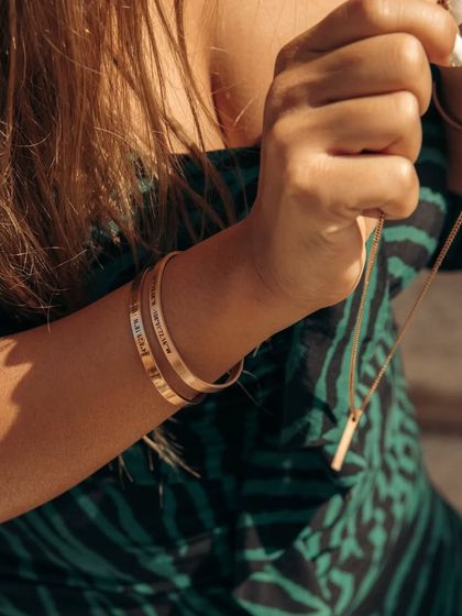 A lifestyle shot showing a woman wearing and holding a set of engraved gold bracelets and a matching bar necklace. This highlights the possibility of creating a full set of custom jewelry.