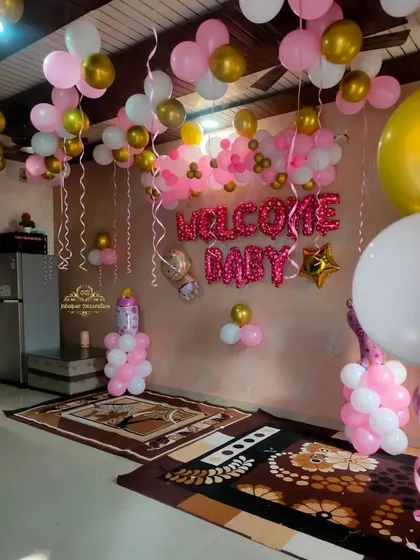 A festive "Welcome Baby" room decoration for a baby girl. The setup includes pink and gold foil letter balloons, ceiling balloons with ribbons, and balloon pillars.