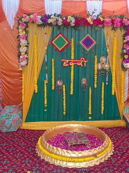 A traditional Haldi ceremony setup with a large brass urli filled with rose petals. The backdrop features green and yellow drapes, marigold garlands, and colorful hangings.