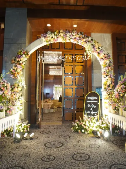 A grand and welcoming entrance for an indoor daytime event. The floral arch, welcome sign, and candle-lined pathway create an impressive first impression for guests.