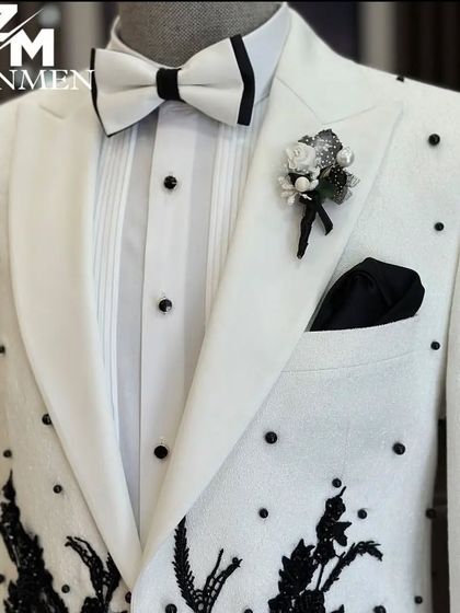 A close-up of the white tuxedo, showing the black pearl studs, the floral lapel pin, and the intricate black embroidery. Every detail is perfectly executed.