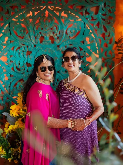 Mother and daughter enjoying the Haldi ceremony. Their coordinated sunglasses add a fun touch to their traditional outfits, a purple bandhani dress for mom and a bright pink ensemble for the bride.
