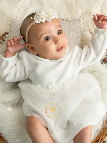 An angelic baby girl in a white outfit lies in a basket, looking up at the camera with curiosity.