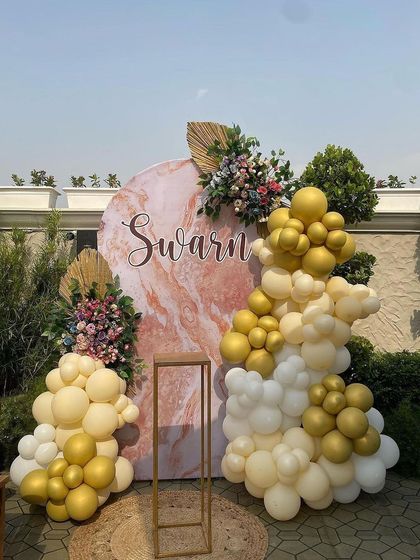 This outdoor baby shower decoration features a beautiful marble-print backdrop personalized with the baby's name. The organic arrangement of cream and gold balloons, paired with floral toppers, creates an elegant and modern rustic feel.