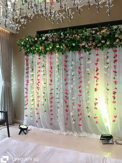 An elegant backdrop for a home party. A sheer white curtain with hanging pink and white flower strings is topped with a lush green and pink floral arrangement.