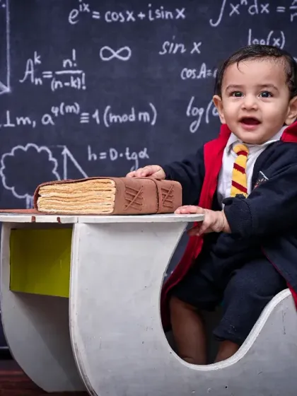 This little wizard is all smiles, dressed in his robes and tie for a magical school-themed photoshoot. The setup with a book-shaped cake on the desk adds a fun, celebratory touch.