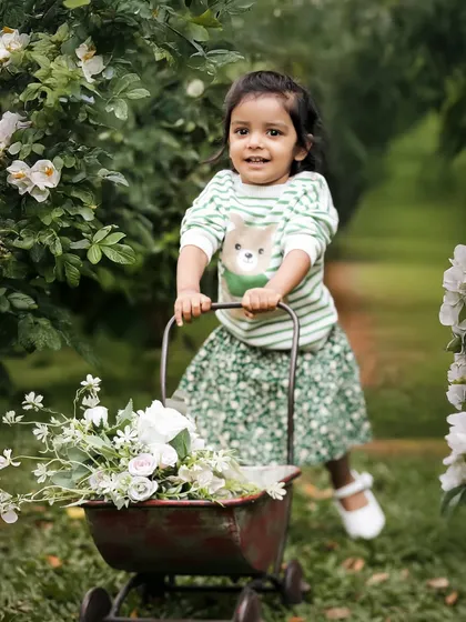 A garden adventure. With her little wagon full of flowers, this toddler is on a mission. The lush green background and her happy expression make this a joyful and vibrant portrait.